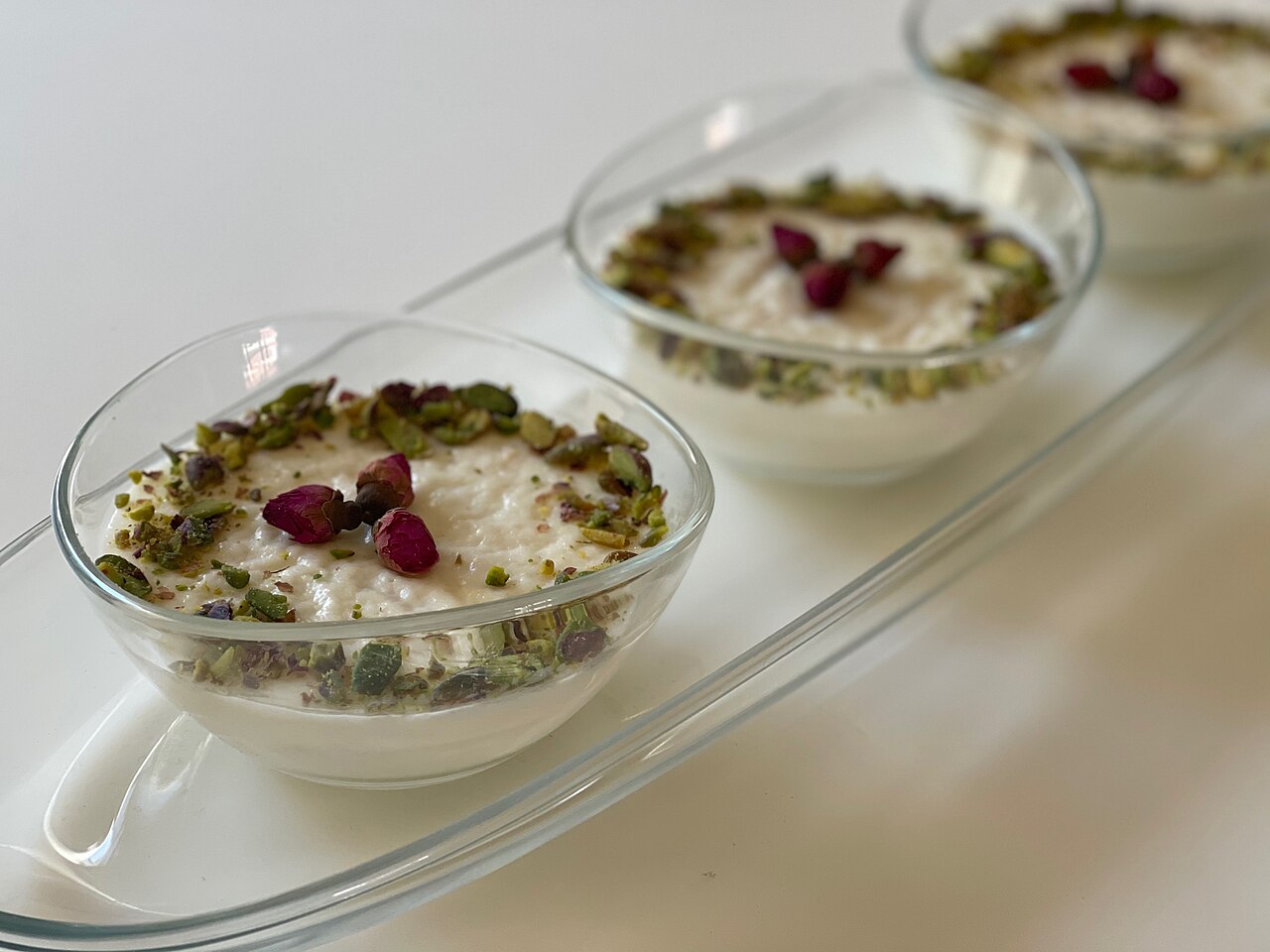 Three glass bowls of white muhallebi topped with pistachio and dried rose petals
